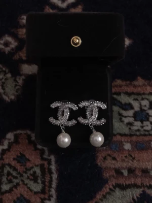 C örhängen med strass och pärla - Snygga C örhängen i klassisk CC-logga, täckta med glittrande strass och med en vacker pärla hängandes. Örhängena är silvriga och kommer i en svart ask. Perfekta för dig som vill ha en lyxig och trendig accessoar.