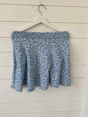 Blommig blå kjol från 157 - Supersöt blå kortkjol från 157 med vitt blommönster och elastisk midja. Kjolen har en lätt utsvängd form och är perfekt för varma dagar. 