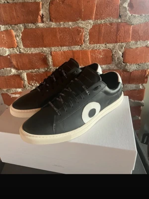 Svarta Oliver Cabell sneakers - Snygga svarta sneakers från Oliver Cabell med vit sula och en stor vit cirkeldetalj på sidan. Skorna har svarta skosnören med märkesnamnet tryckt i vitt och är tillverkade i slätt läder. Perfekta för dig som gillar stilrena och moderna sneakers.