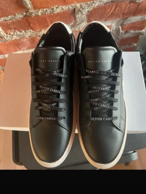 Svarta sneakers från Oliver Cabell - Stilrena svarta sneakers i läder från Oliver Cabell med vit sula och vit cirkeldetalj på sidan. Skorna har svarta skosnören med varumärket tryckt i vitt och diskret logga på plösen. Perfekta för en trendig och clean look.