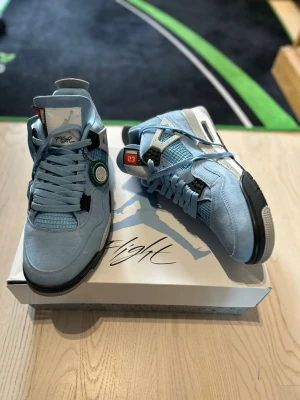 Air Jordan 4 Retro ”University Blue” Nyskick! - Säljer ett par extremt fräscha och 100% äkta Air Jordan 4 Retro i den ikoniska färgställningen ”University Blue” helt nya. Modellen kännetecknas av sin premiummocka i ljusblått, kombinerat med de klassiska cementgrå detaljerna med svarta stänk. Skorna köptes ursprungligen via StockX, och den gröna ”Verified Authentic”-taggen sitter fortfarande kvar på skon (QR-kod på baksidan).  Detaljer:  Modell: Air Jordan 4 Retro (CT8527-400)  Färg: University Blue/Black/Tech Grey  Storlek: EU 46 / US 12 / UK