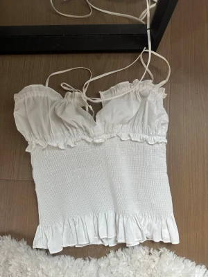 Vit smockad topp med volang - Söt vit topp med smockad midja, tunna axelband och volangdetaljer upptill och nedtill. Toppen har en liten knytning framtill och är ärmlös, perfekt för en trendig och luftig look. Aldrig använt köpt förra året på zara💘