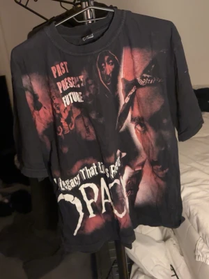 Svart Tupac t-shirt med tryck - Cool svart t-shirt med stort Tupac-tryck fram och bak. Texten 'Past Present Future' och 'A Legacy That Lives Forever' syns tillsammans med flera porträtt av Tupac i rött och vitt. Perfekt för dig som gillar streetwear och hiphop.