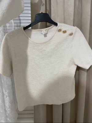 Krämvit topp med guldknappar H&M - Superfin krämvit topp från H&M med kort ärm och tre dekorativa guldknappar på axeln. Tjockare, lite texturerat material som känns lyxigt och trendigt. Perfekt att styla med jeans eller kjol för en clean look.
