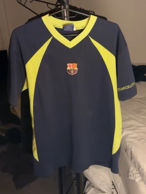Retro FC Barcelona träningströja  - Snygg träningströja från FC Barcelona i mörkblått med neongula detaljer och klubbmärke på bröstet. Tröjan har korta ärmar, v-ringad krage och är tillverkad i ett ventilerande polyestermaterial. Perfekt för fotbollsträning eller gymmet.