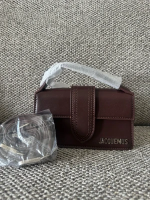 Mörkbrun Jacquemus handväska - Säljer en stilren mörkbrun handväska från Jacquemus i slätt läder. Väskan har ett elegant lock med bred rem och silverfärgad Jacquemus-logga framtill. Medföljer matchande axelrem. Perfekt för dig som gillar trendiga accessoarer.made in italy,dustbag kommer