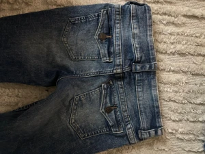 Blå bootcut jeans från H&M - Säljer ett par blå bootcut jeans från H&M med låg midja och justerbar linning. Jeansen har klassiska bakfickor med knappdetaljer och slitna tvättade partier för en cool look.