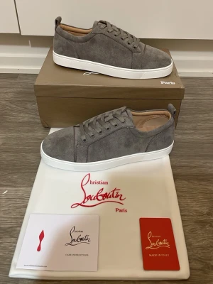 Christian Louboutin sneakers - Stilrena gråa sneakers i mocka från Christian Louboutin med vita sulor och klassisk låg profil. Skorna har matchande gråa skosnören och diskreta detaljer, perfekt för en clean och modern look. Tillverkade i Italien och levereras med originalkartong och dustbag.