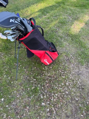 Golfset med TaylorMade väska - Golfset med svart och röd TaylorMade bärbag, flera golfklubbor med skydd, bland annat King och R15. Väskan har flera fack och praktiskt bärhandtag. Perfekt för golfbanan och enkel att ta med tack vare stativ och axelrem.