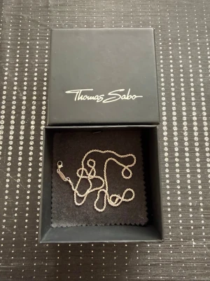 Silverhalsband 925 från Thomas Sabo - Säljer ett stilrent silverhalsband från Thomas Sabo. Kedjan är tunn och elegant med klassisk karbinhake och märkets logga ingraverad på brickan vid låset. Perfekt för att bära som det är eller kombinera med berlocker. Nypris för 800kr