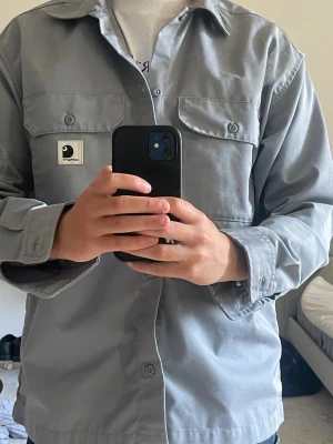 Ljus blå overshirt från Carhartt WIP - Snygg ljusblå overshirt från Carhartt WIP med klassisk krage och två bröstfickor med lock och knappar. Overshirten har raka ärmar och en stilren look med Carhartt-logga på vänster ficka. Perfekt att bära öppen eller stängd över en t-shirt.