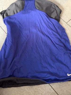 Nike Dri-Fit blå träningslinne - Snyggt blått träningslinne från Nike med svarta detaljer och Dri-Fit-material som andas. Perfekt för gymmet eller löpning. Klassisk racerback-modell och liten vit Nike-logga framtill.