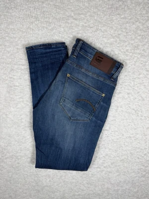 Blå slim fit jeans från G-Star RAW - Snygga blå slim fit jeans från G-Star RAW med klassisk femficksdesign och läderpatch bak i midjan. Jeansen har en lätt tvättad look och smal passform som sitter tajt längs benen. Perfekta för en trendig och avslappnad stil.