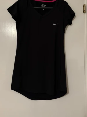 Svart Nike Dri-Fit sporttröja - Svart sporttröja från Nike med Dri-Fit-teknologi som transporterar bort fukt. Tröjan har korta ärmar, V-ringning och en liten vit Nike-logga på bröstet, den är som en tunika i längden. Perfekt för träning och sportaktiviteter.