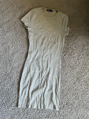 Grå ribbad klänning från FB Sister - En enkel och stilren grå ribbad klänning från FB Sister. Modellen är kroppsnära med korta ärmar och rund halsringning. Perfekt för dig som gillar en minimalistisk look och vill ha något bekvämt och snyggt.