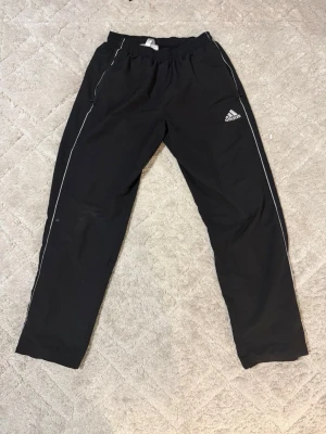 Svarta mjukisbyxor från Adidas - Svarta mjukisbyxor från Adidas med vita detaljer längs sidorna och broderad logga på vänster ben. Byxorna har resår i midjan och dragkedjeförsedda fickor. Perfekta för chill eller träning.