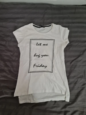 Vit t-shirt med text från Tiki - Vit t-shirt från Tiki med svart text på framsidan där det står 'Let me hug you Friday' i en rektangel. T-shirten har rund halsringning och korta ärmar. Perfekt för en chill och avslappnad stil.