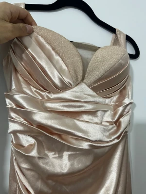 Champagnefärgad långklänning i satin - Elegant långklänning i glansig champagnefärgad satin med vadderad byst och draperade detaljer. Klänningen har breda axelband och en figurnära passform som ger en lyxig känsla. Perfekt för speciella tillfällen när du vill sticka ut.