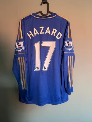 Chelsea Hazard 17 långärmad tröja - Blå långärmad fotbollströja från Chelsea FC med Hazard och nummer 17 på ryggen. Tröjan har gula ränder på ärmarna och Barclays Premier League-patch på båda ärmarna. Perfekt för fans av engelsk fotboll och lira i!