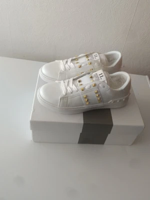 Valentino Rockstud - Snygga vita sneakers Valentino Garavani med guldfärgade nitar längs sidorna och klassisk låg profil. Skorna har en stilren design med vita snören och detaljer i läder. Perfekta för dig som vill sticka ut med en lyxig touch.