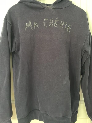 Mörkblå hoodie med strass från Gina Tricot - Cool mörkblå hoodie från Gina Tricot med texten 'MA CHÉRIE' i strass på bröstet. Klassisk modell med huva och ribbade muddar. Perfekt för en avslappnad och trendig look.