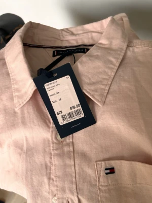 Ljusrosa skjorta från Tommy Hilfiger - Snygg ljusrosa skjorta från Tommy Hilfiger med klassisk krage och liten broderad logga på bröstfickan. Skjortan är i ett mjukt bomullstyg och har knappar framtill. Perfekt för en stilren look.