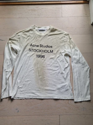 Vit långärmad topp Acne Studios - Vit långärmad topp från Acne Studios med tryck 'Acne Studios STOCKHOLM 1996' på bröstet. Klassisk rund halsringning och avslappnad passform. Perfekt för en clean och stilren look.