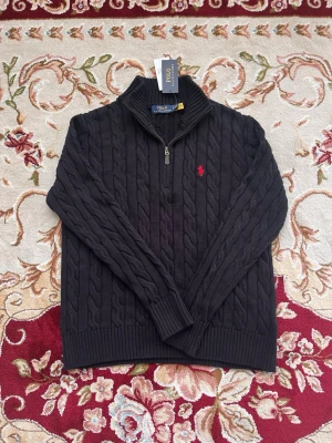 Mörkgrå kabelstickad tröja Polo Ralph Lauren - Mörkgrå kabelstickad tröja från Polo Ralph Lauren med halv dragkedja och hög krage. Tröjan har ribbstickade muddar och nederkant samt den klassiska röda logotypen broderad på bröstet. Perfekt för lager-på-lager och snygg till jeans.