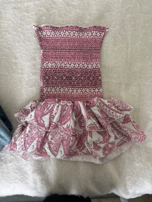 Rosa och vit volangklänning - Supersöt kort klänning med smockad överdel i rosa och vitt samt volangig kjol med blommigt mönster. Ärmlös och axelbandslös modell, perfekt för varma dagar. Materialet känns lätt och luftigt. 
