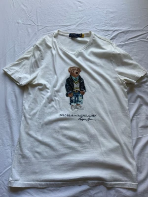 Vit Polo Bear t-shirt från Ralph Lauren - Vit t-shirt från Ralph Lauren med ikoniskt Polo Bear-tryck på bröstet. Trycket visar en nalle i preppy outfit och texten 'POLO BEAR by RALPH LAUREN'. Kan gå ner i pris vid snabb affär! Passar L men även M. Varan är äkta!