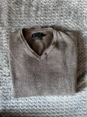 Beige V-ringad stickad tröja Riley - Snygg beige stickad tröja från Riley med klassisk V-ringning. Tröjan har långa ärmar och ribbade muddar vid ärmslut och nederkant. Perfekt att matcha med skjorta eller t-shirt för en clean look.