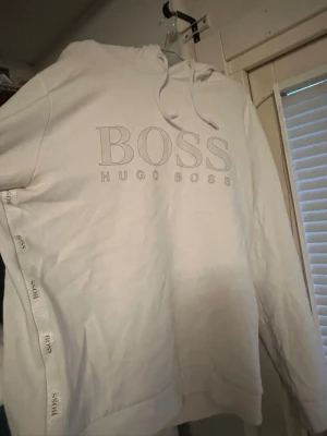 Vit hoodie från Hugo Boss - Snygg vit hoodie från Hugo Boss med stor BOSS-logga framtill och diskreta logoband längs sidorna. Hoodien har dragsko i huvan och en clean, stilren look som passar perfekt till streetwear eller chill vardagsstil.