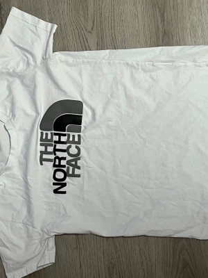 Vit t-shirt från The North Face - Snygg vit t-shirt från The North Face med stor svart logga på framsidan. Klassisk passform och rund halsringning, perfekt för en clean och stilren look. Tillverkad i mjuk bomull som känns skön mot huden.