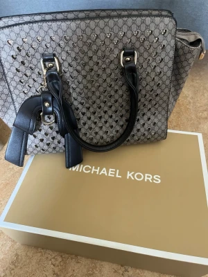 Michael kors väska - Snygg grå handväska från Michael Kors med klassiskt MK-mönster och svarta handtag i läder. Använt 2 gånger. Kvitto finns även.