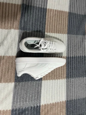 Vita Puma sneakers i läder - Fräscha helvita sneakers från Puma med klassisk låg siluett. Skorna har ovandel i slätt läder, perforerad tå för extra ventilation och diskreta detaljer längs sidorna. Perfekta för en clean och stilren look.