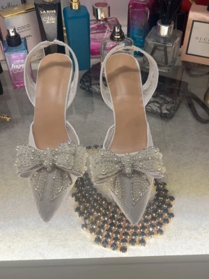 Vita spetsiga pumps med pärlrosett - Snygga vita pumps med spetsig tå och hög klack. Skorna har en stor dekorativ rosett framtill täckt av pärlor och meshdetaljer längs sidorna. Perfekta för dig som vill sticka ut med en elegant och trendig look.