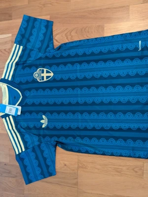 Blå Sverige fotbollströja Adidas. Storlek-.L - Helt ny  blå fotbollströja från Adidas med Sveriges landslagsemblem på bröstet. Tröjan har ett mönster i olika blå nyanser, gula detaljer vid krage och axlar samt Adidas-logga. Perfekt för dig som vill stötta Sverige med stil.