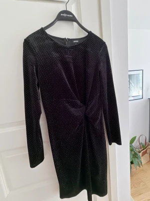 Svart glittrig sammetsklänning - Snygg svart sammetsklänning från Daily Story med glittriga prickar, lång ärm och twistad detalj framtill. Kort modell som passar perfekt när du vill sticka ut lite extra. Dragkedja i ryggen för enkel påklädning.