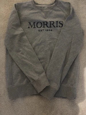 Grå sweatshirt från Morris - Snygg grå sweatshirt från Morris med tryckt logga och text 'EST 1968' på bröstet. Tröjan har rund halsringning, ribbade muddar vid ärmslut och nederkant samt en klassisk relaxed fit. Perfekt för en chill och stilren look.