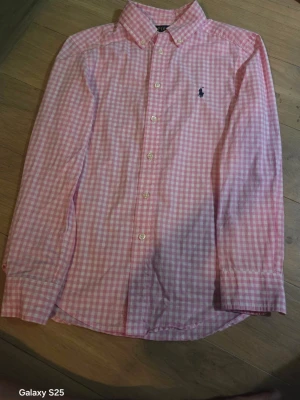 Rosa rutig skjorta från Polo Ralph Lauren - Klassisk rosa och vit rutig skjorta från Polo Ralph Lauren med broderad logga på bröstet. Skjortan har lång ärm, knappar framtill och krage. Perfekt för dig som gillar preppy stil och vill ha något som sticker ut lite extra.