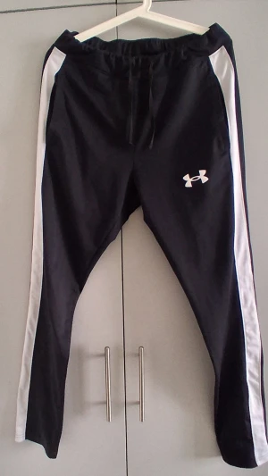 Svart set från Under Armour - Säljer ett svart träningsset från Under Armour med vita detaljer längs ärmar och ben. Setet består av en zip-jacka och matchande byxor, båda med logga på bröst och lår. Perfekt för gymmet eller löprundan. Materialet är mjukt och stretchigt för maximal komfort.