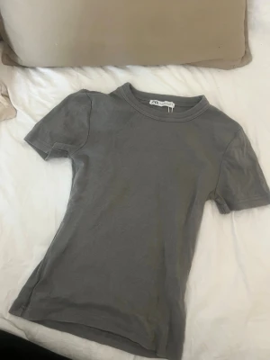 Grå ribbad t-shirt från Zara - Säljer en enkel och stilren grå t-shirt från Zara. T-shirten är ribbad, har korta ärmar och en klassisk rund halsringning. Perfekt basplagg som passar till allt och är superbekväm tack vare det mjuka bomullsmaterialet.