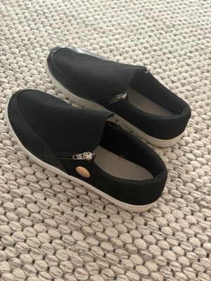 Svarta slip-on sneakers med dragkedja - Svarta sneakers i slip-on-modell med dragkedja på sidan och vit sula. Ovandelen är i textil och detaljer i syntetmaterial. Snygg och enkel design som passar till det mesta.
