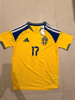 Svensk landslagströja Adidas gul - Gul svensk landslagströja med blå detaljer och nummer 17 och gyökeres på ryggen. Tröjan har korta ärmar, blått och vitt mönster på axlarna . Tillverkad i lätt och funktionellt material.