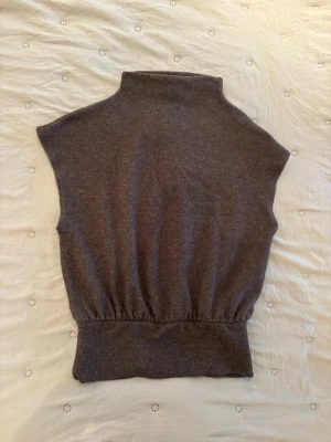 Brun ärmlös stickad topp Gina Tricot - Säljer en brun ärmlös stickad topp från Gina Tricot med hög krage och bred ribbad kant nedtill. Perfekt att styla över en skjorta eller t-shirt för en trendig lager-på-lager-look. Mjuk och skön kvalitet.