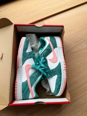 Nike Dunk Low ”Washed Teal, Bleached Coral” - Nike Dunk Low sneakers i vitt, grönt och rosa med ormmönstrade detaljer. Skorna har turkosa skosnören, rosa swoosh och sula samt perforerad tåbox för extra ventilation. Klassisk låg siluett och ikonisk Nike-logga på plösen.