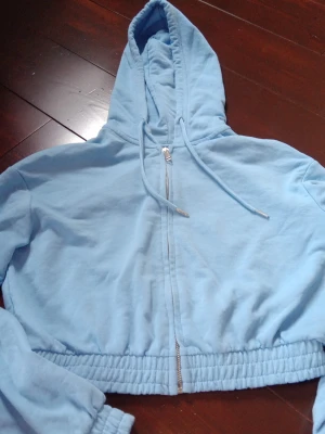 Ljusblå croppad hoodie med dragkedja - Snygg ljusblå croppad hoodie med dragkedja framtill och justerbar huva med snören. Mjuk resår i midjan och vid ärmsluten för en skön passform. Perfekt för en avslappnad och trendig look. Materialet känns som bomullsmix.