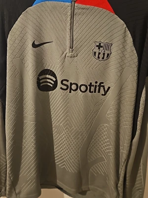 Grå FC Barcelona träningströja Nike - Snygg grå träningströja från Nike med FC Barcelona-logga och Spotify som sponsortryck. Tröjan har halv dragkedja, diskret mönster i tyget och detaljer i blått och rött vid kragen. Perfekt för fotbollsträning eller som sportig streetwear. Säljer den för den kommer inte till användning 