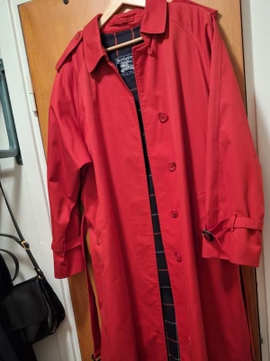 Röd trenchcoat från Burberry - Snygg röd trenchcoat från Burberry med klassisk krage, knäppning framtill och bälte i midjan. Jackan har axelklaffar, justerbara ärmslut och ett rutigt innerfoder. Perfekt för dig som vill sticka ut med en ikonisk och tidlös look.
