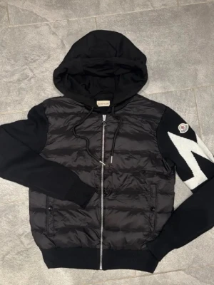Svart hoodie från Moncler med dragkedja - Snygg svart hoodie från Moncler med vadderad framsida, stickade ärmar och stor huva. Passar perfekt för kommande sommarkvällarna 